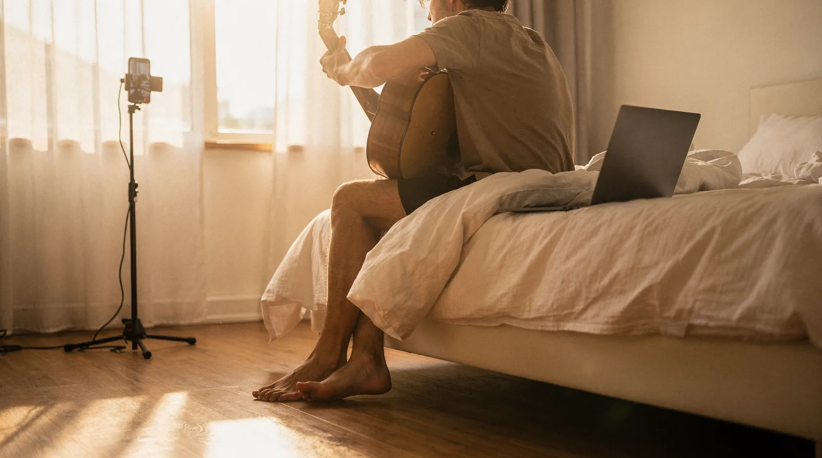 A songwriter on the edge of a bed with a guitar, laptop and phone tripod in morning light